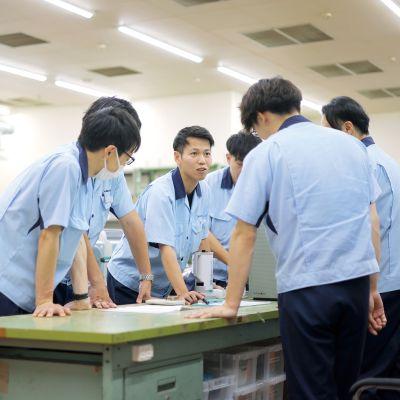 ダイニチ工業の開発現場で、図面や資料を囲んで打ち合わせを行う技術職の社員たち。複数の若手社員が意見を出し合い、チームで製品開発に取り組む様子。