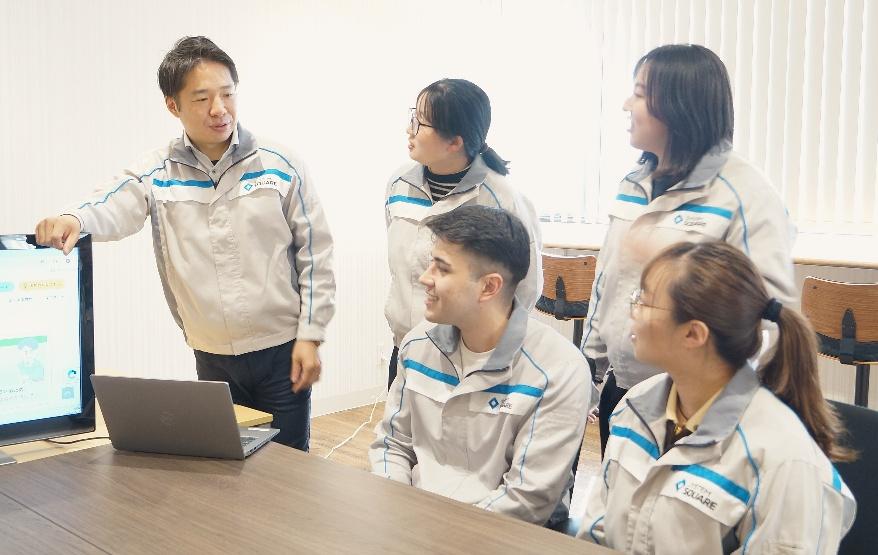 新潟県長岡市にある異物検査機メーカー、株式会社システムスクエアの社内打ち合わせ風景。お揃いの作業服を着た社員たちが笑顔で、モニターとノートパソコンを前に談笑している。機械・電気・ソフトの各専門分野が連携し、人々の安心安全を守る新技術開発に挑む。
