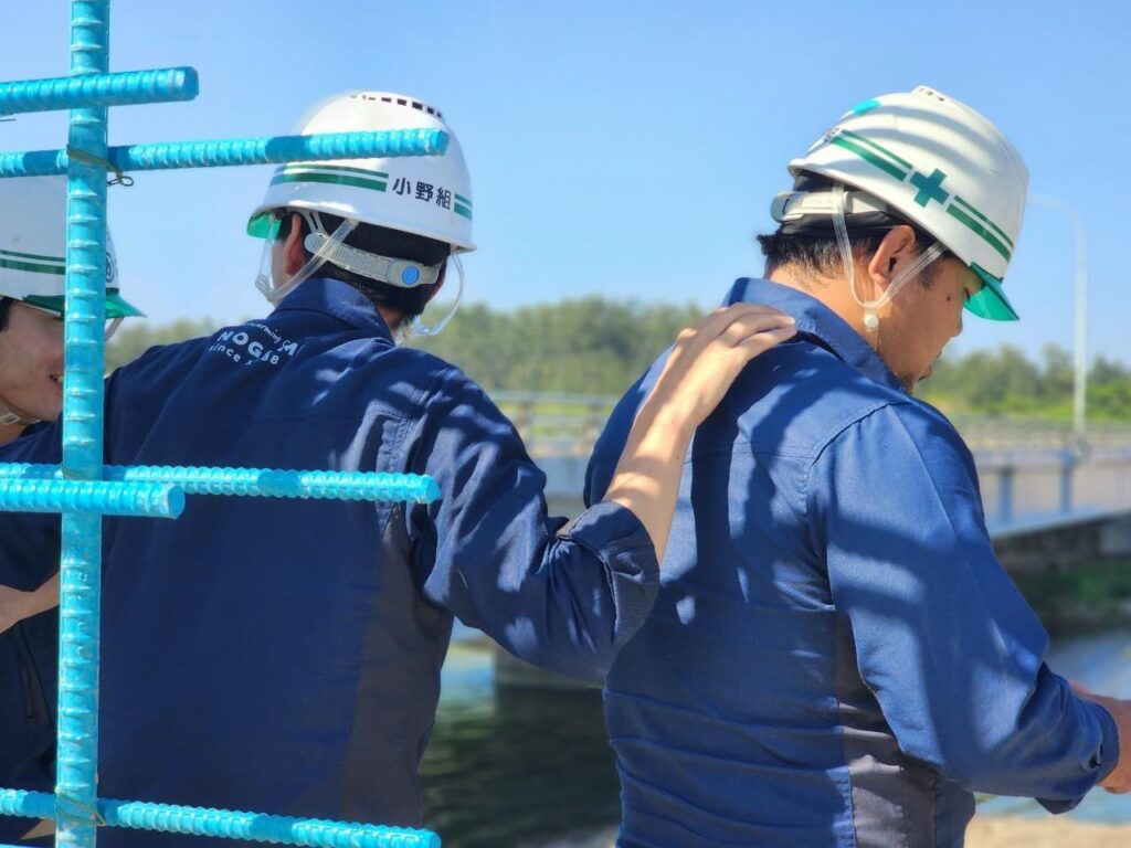 建設現場での活気あるチームワーク。小野組の作業服を着たスタッフが談笑する、風通しの良い職場環境のイメージ。