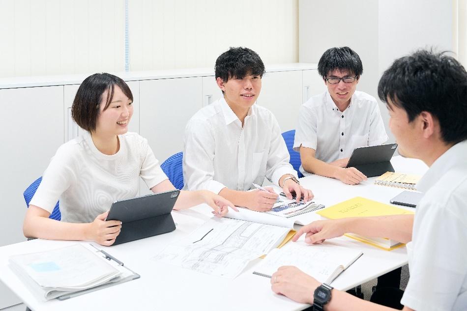 株式会社ネクスコ・エンジニアリング新潟の社内でのミーティング風景。先輩社員の指導を受けながら、図面やタブレットを使って和やかに話し合う若い社員たち。