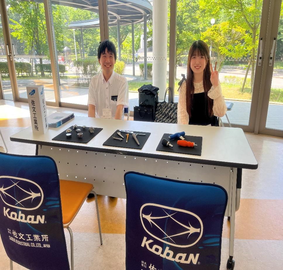 新潟市江南区に本社を置く株式会社佐文工業所の採用イベントや展示会の様子。精密加工技術を活かしたミシン部品や医療機器などの製品サンプルを前に、若手社員が笑顔で対応する。ボトムアップな社風と温かい企業文化が伝わる、地域に根ざしたものづくり企業の紹介画像。