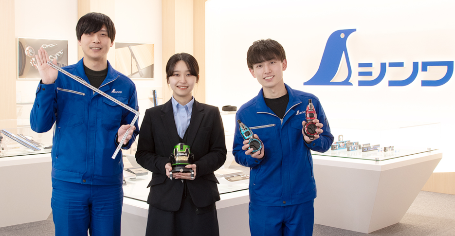 シンワ測定株式会社の社員3名が、新潟県燕三条発の測定機器を手に笑顔で並ぶ写真。左の男性は曲尺、中央の女性はレーザー墨出し器、右の男性はハンディ墨つぼを持ち、背景にはペンギンマークの企業ロゴ。