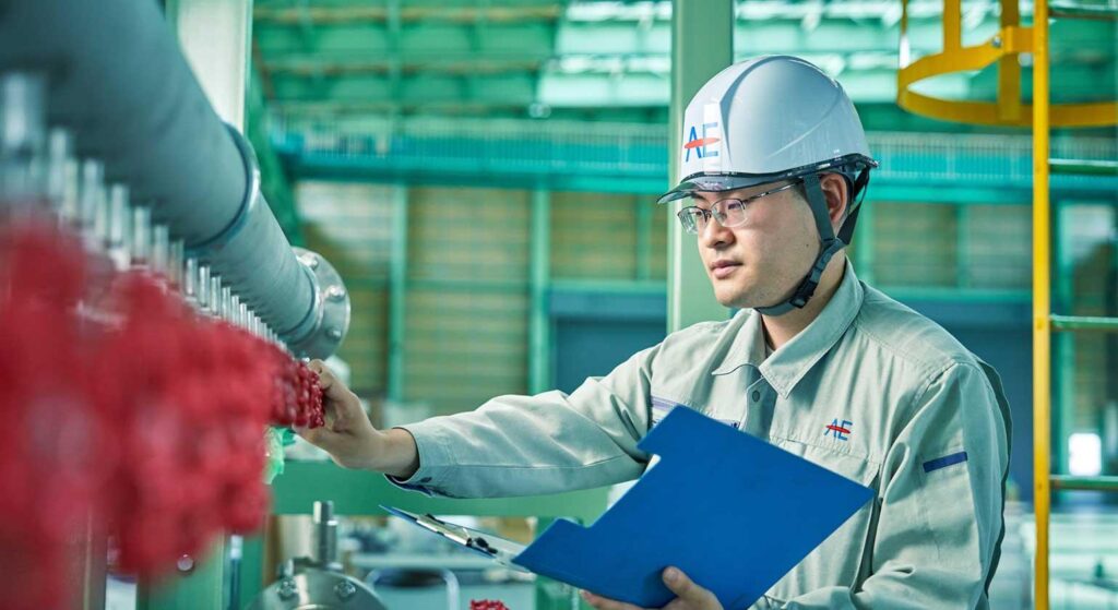 アドバンエンジ株式会社の製造現場にて、ヘルメットと作業着を着用した技術職社員が工業炉の設備点検を行っている様子。新潟市にある同社の工場内にて、クリップボードを持ちながら機械の調整箇所を確認する。オーダーメイドの大型設備のメンテナンス業務の光景。