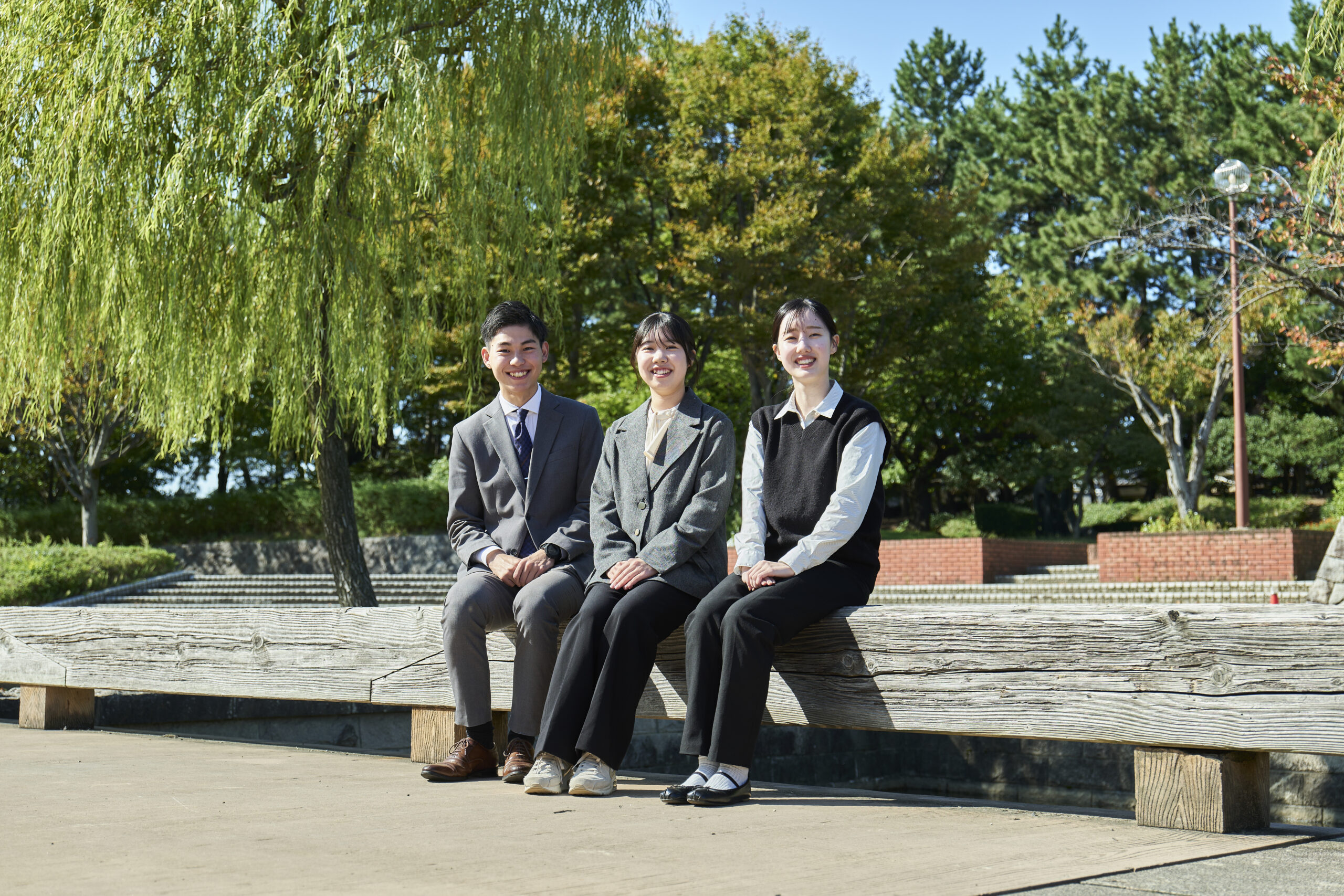 新潟市中央区の公園で、笑顔で木製ベンチに座るNSGグループの若手社員3名。教育や医療・福祉など101法人を展開するNSGグループでは、新潟から「世界一豊かで幸せなまち」を創る志を持つ人材を募集しており、地方創生の未来を担う新卒総合職の爽やかなイメージを表現。