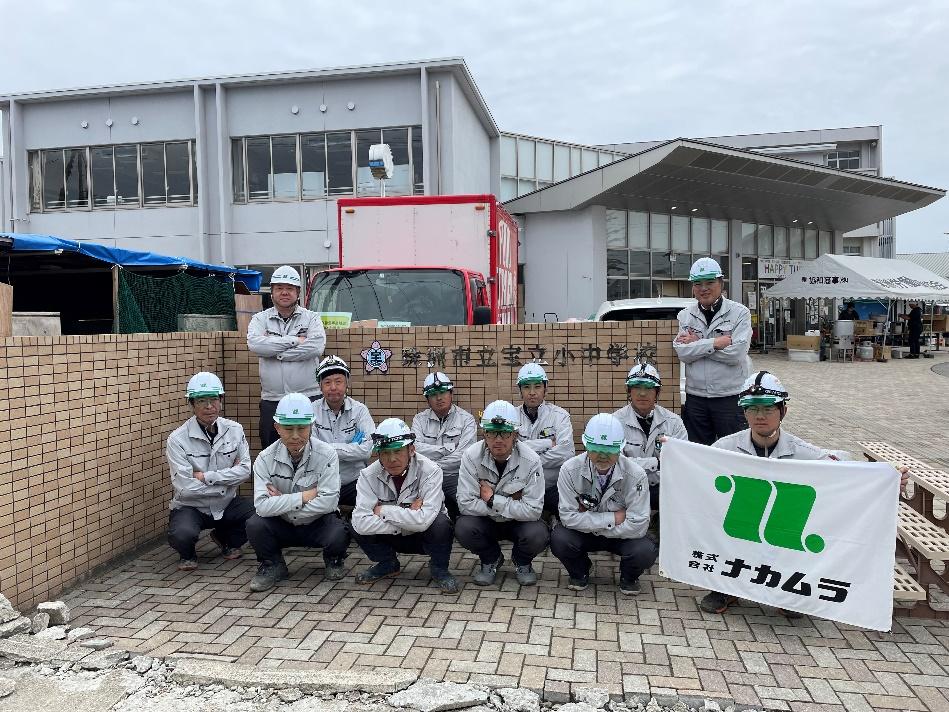 能登半島地震の支援活動に従事する株式会社ナカムラの作業員たち。珠洲市立宝立小中学校の校舎と赤色のトラックを背景にした集合写真